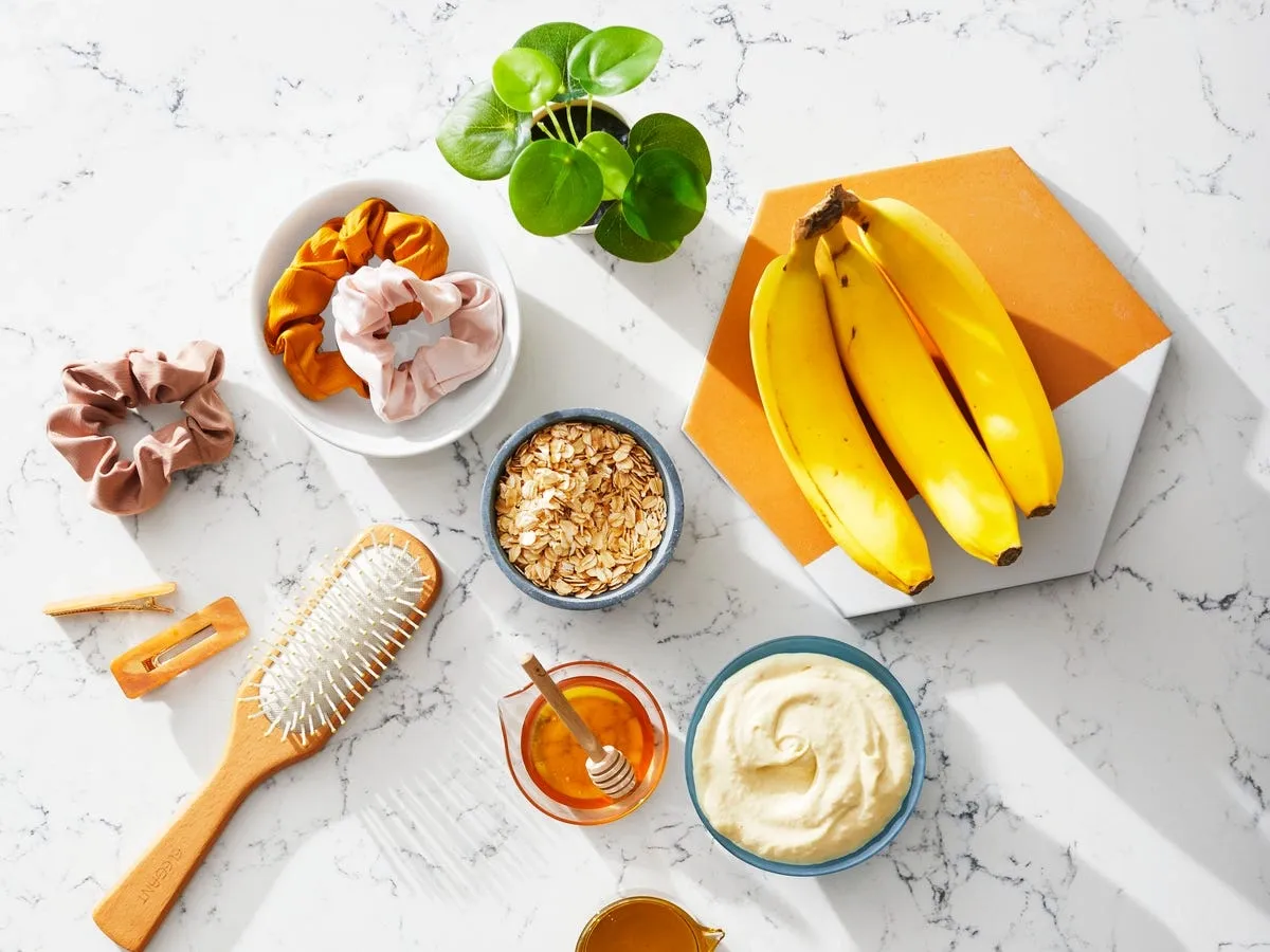 Flat lay of natural ingredients and tools for DIY hair masks, including bananas, oats, honey, cream, and hair accessories on marble.
